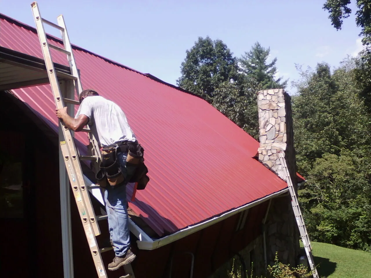 Roofer on a ladder beside a red metal roof and stone chimney — Soffit & Fascia Repair by Roofing Brothers in Skykomish